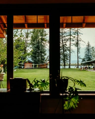 View through large cabin windows across a green lawn to a lake and outbuildings, with houseplants on the sill